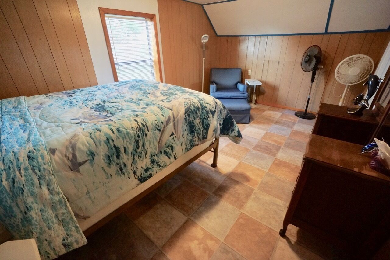 A bedroom with a bed, a chair, a desk, and two fans.
