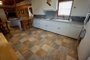 A kitchen with a tiled floor, white cabinets, a dining area with chairs, and a television in the background.