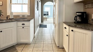 Une cuisine avec des meubles blancs, un comptoir en granit et un sol carrelé. Il y a une fenêtre sur la gauche et une porte qui donne sur une autre pièce.