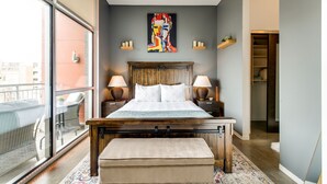 Quarto com uma janela ampla, cama de madeira, duas luminárias de cabeceira e um quadro colorido na parede.