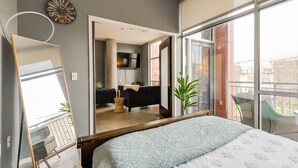 Quarto moderno com cama grande, espelho, relógio e vista para uma sala de estar com sofá e televisão.