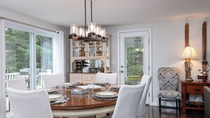A dining area with a round table, white upholstered chairs, a chandelier, and a view of the outdoors through large windows.