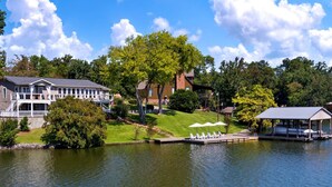 Une propriété au bord du lac comprenant une grande maison, un bâtiment plus petit et un quai équipé de chaises longues.