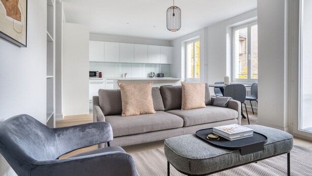 A modern living room with a grey sofa, a grey armchair, and a coffee table. There is a kitchen in the background with white cabinets and a dining area with a table and chairs. - Airy Vorstädte 3.5 Room w/ W/D & Storage, 2 min to Market, by Blueground (Basel)