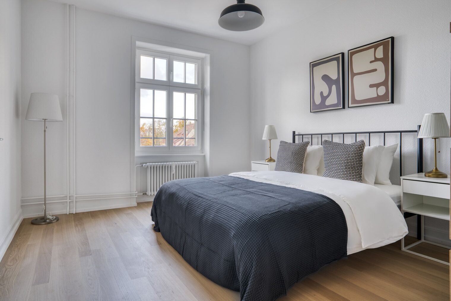 Een slaapkamer met een bed, nachtkastjes, een vloerlamp, een raam en abstracte wanddecoratie.