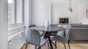 A modern living room with a round dining table, grey upholstered chairs, a white vase, a television, and a sofa. - Airy Vorstädte 3.5 Room w/ W/D & Storage, 2 min to Market, by Blueground (Basel)