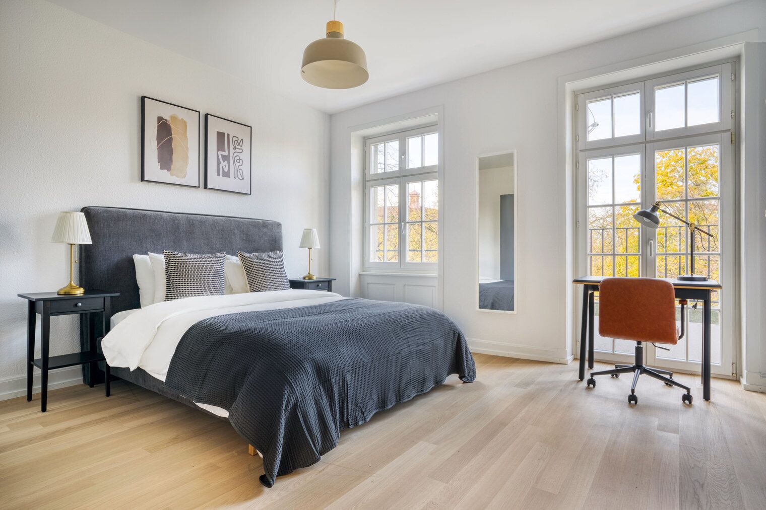 Un dormitorio moderno con una cama grande, dos mesitas de noche, un escritorio con silla y ventanas amplias con follaje otoñal en el exterior.
