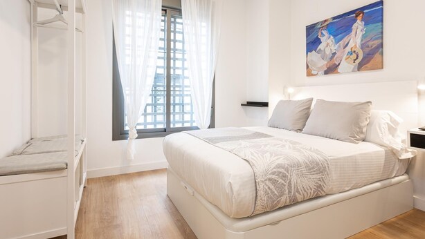 Un dormitorio con cama, mesita de noche, una silla, ventana con cortinas y un cuadro en la pared.
