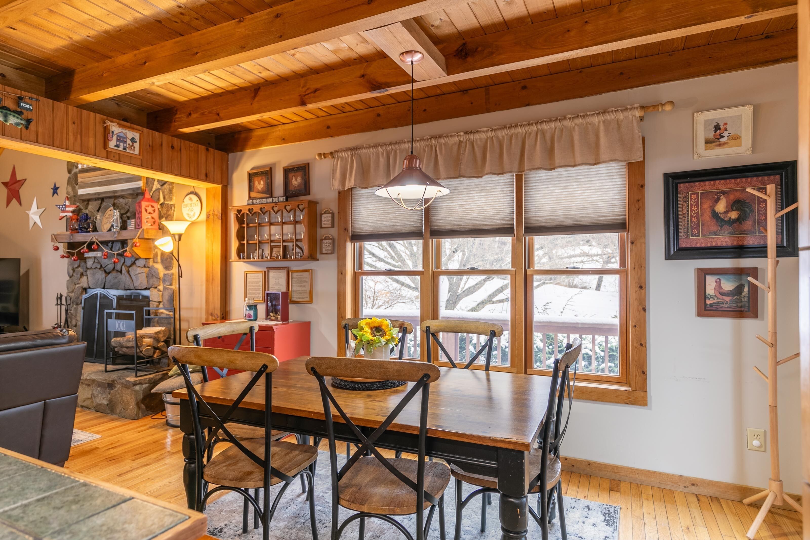 A cozy dining area with a wooden table and chairs, a stone fireplace, and a large window with blinds.