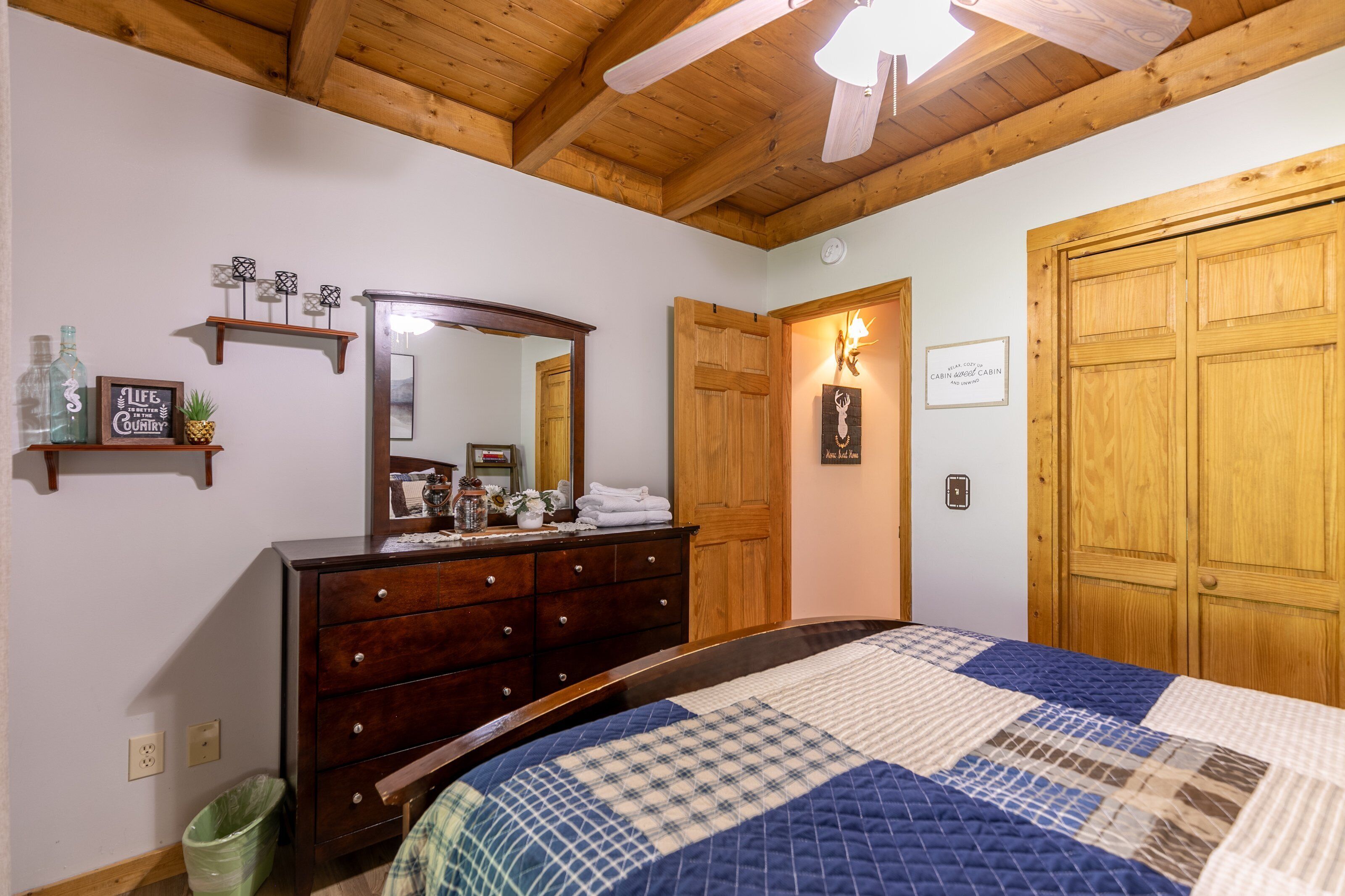 A bedroom with a wooden ceiling, a large bed with a quilt, a dresser, a mirror, and wooden doors.