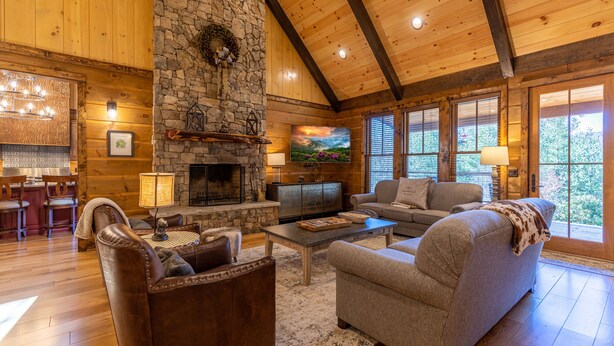 A cozy living room with a stone fireplace, wooden ceiling beams, and large windows. - Papa's View - Pet Friendly Luxury Log Cabin in Valle Crucis with Views and Hot Tub! (Banner Elk)