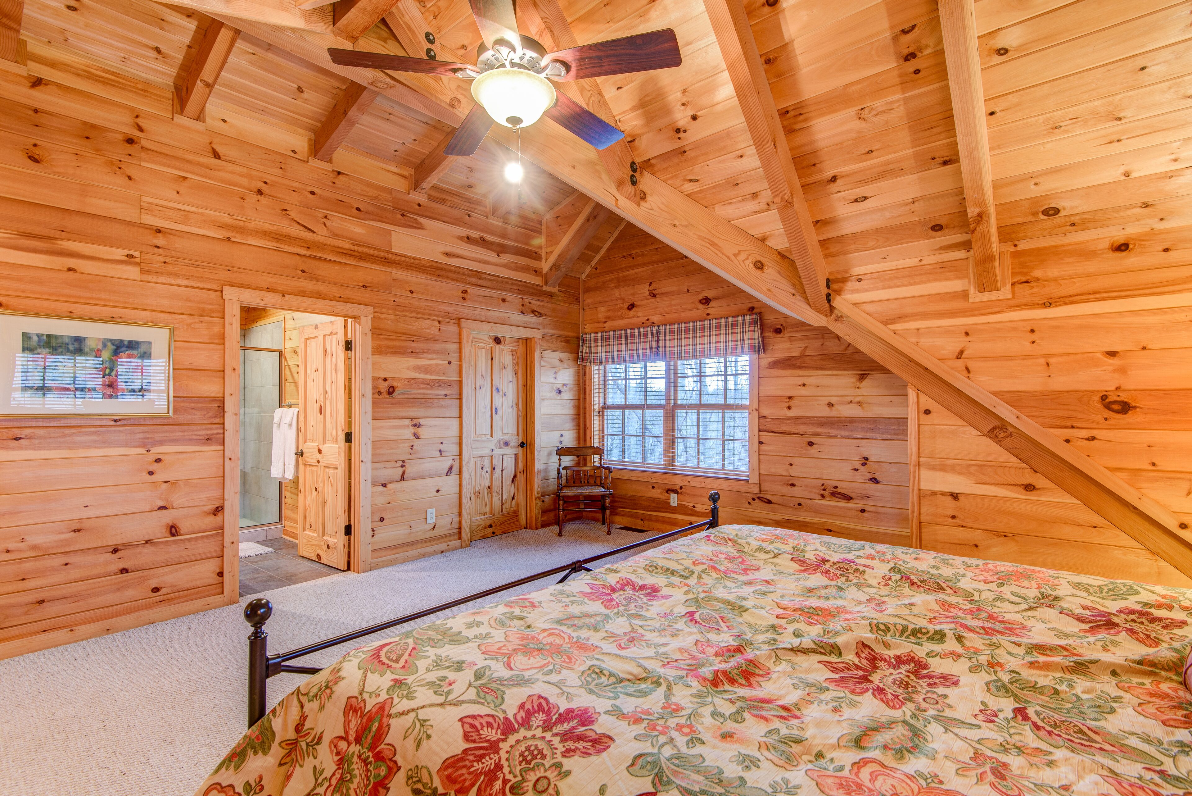 Une chambre dans une cabane en bois, avec un lit, un ventilateur de plafond et une fenêtre agrémentée d’une lambrequin à carreaux.