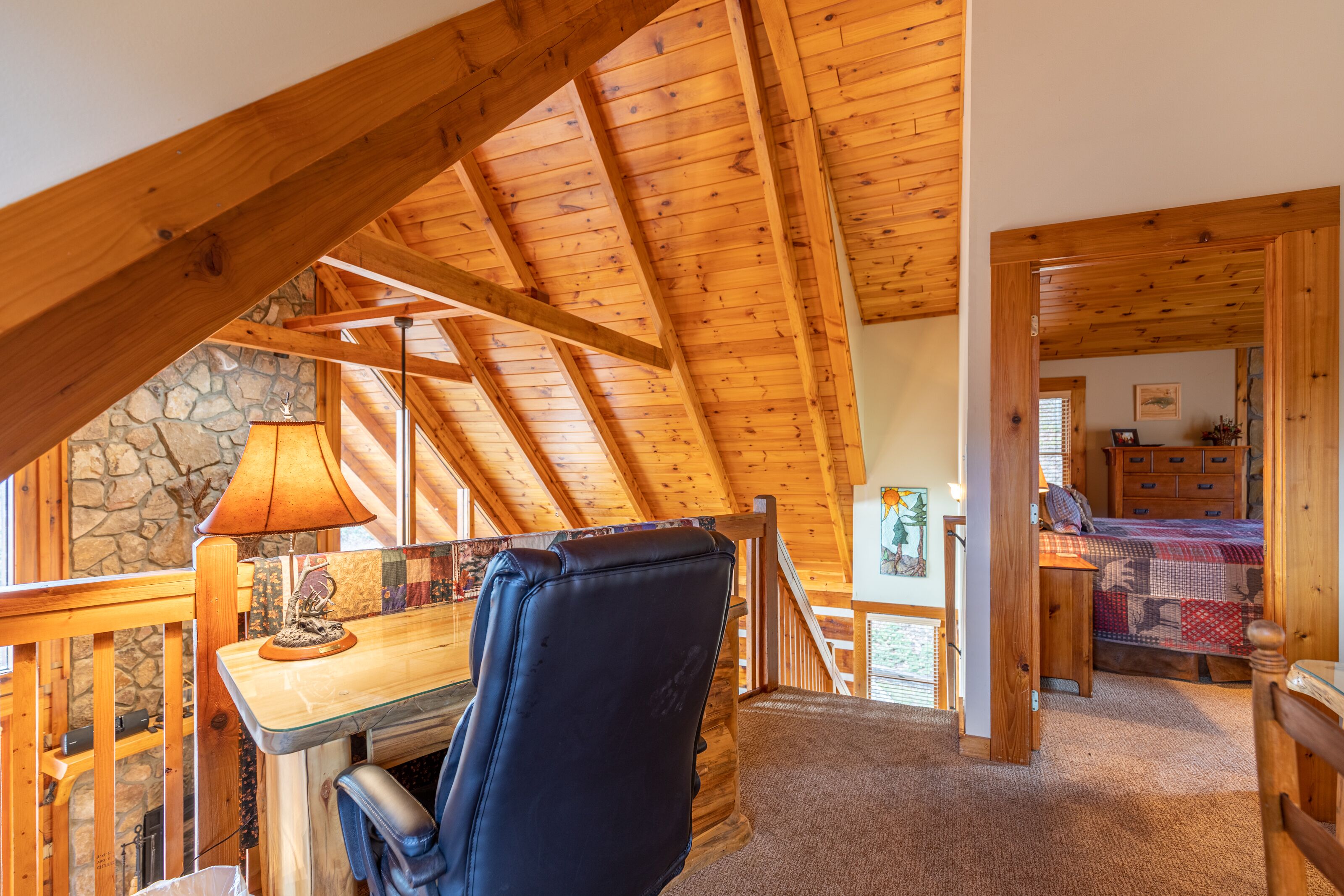 A cozy attic bedroom with a wooden ceiling, a stone wall, a desk with a lamp, a chair, and a bed with a quilt.