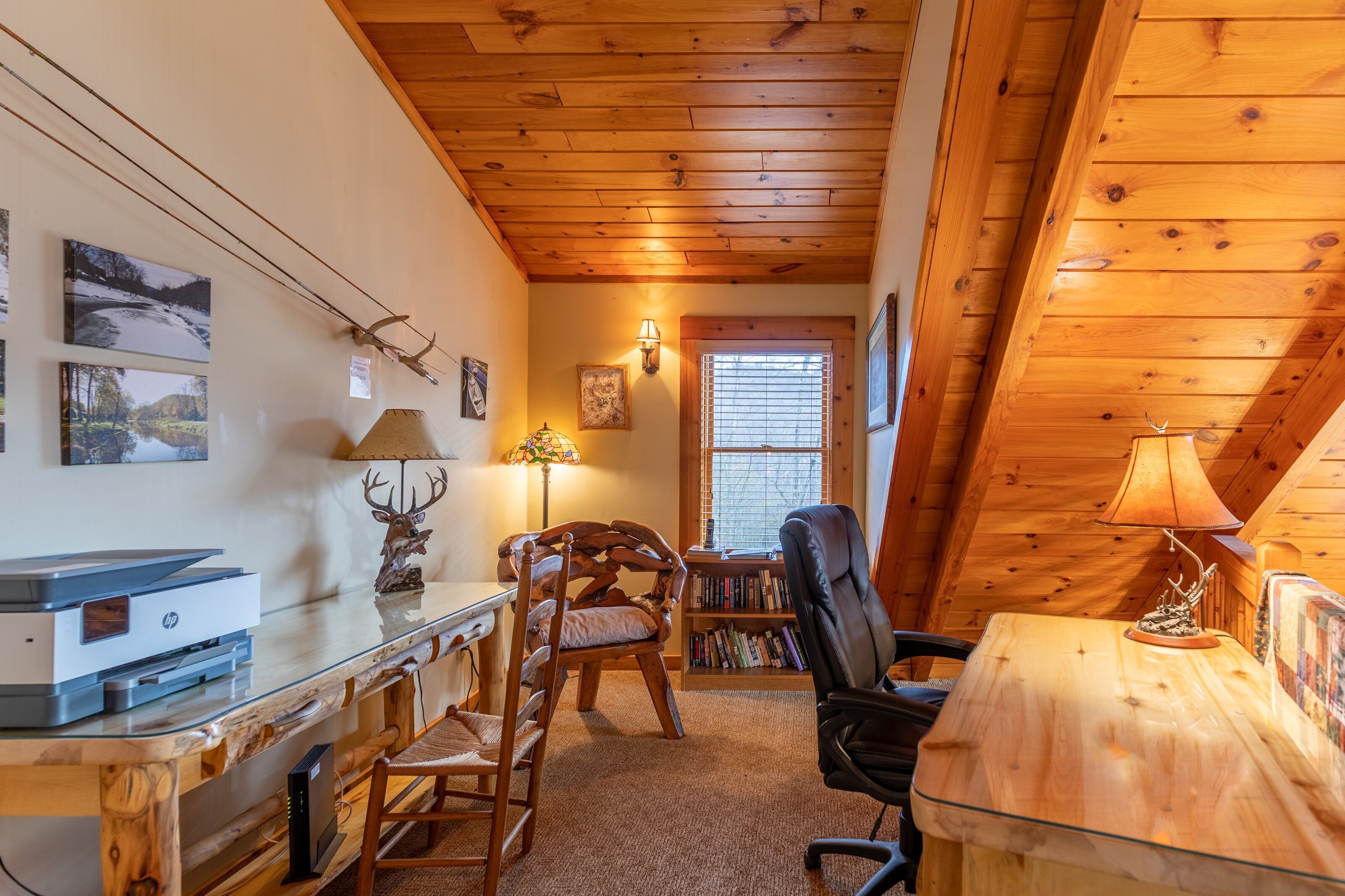 A cozy cabin interior with a wooden ceiling, a desk with a printer, a chair, a bookshelf, and a lamp.