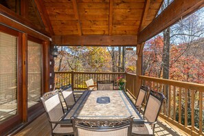 A wooden deck with a table and chairs, surrounded by a wooden railing, overlooking a forest with autumn foliage. - Ambleside- Pet friendly, 3 fireplaces, game room, views, lake fishing access (Vilas)