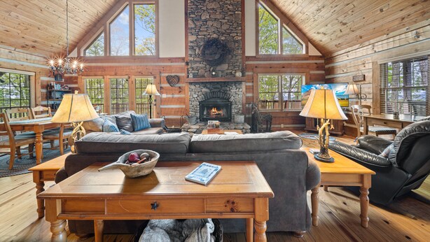 Interior de una cabaña de troncos con chimenea de piedra, muebles de madera y ventanas amplias.
