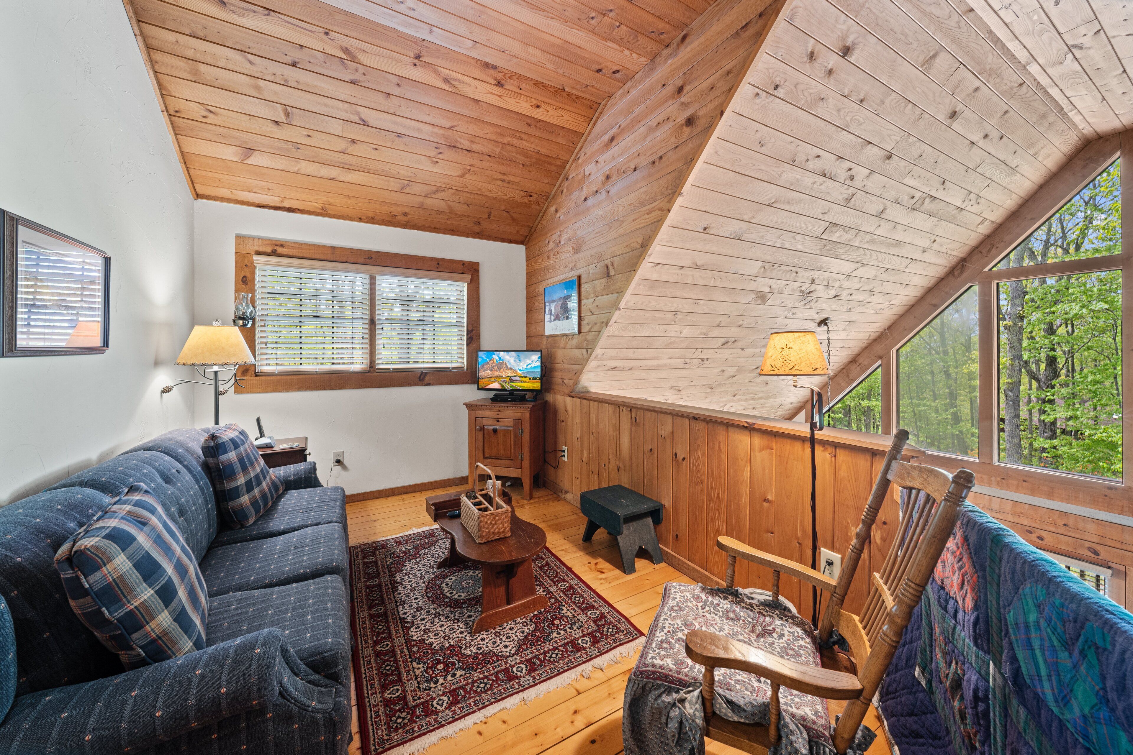 A cozy cabin interior with a wooden ceiling, a sofa, a small table, a chair, and a window offering a view of the forest.