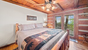 Un dormitorio de cabaña de madera con una cama grande, un ventilador de techo y una ventana con persianas.