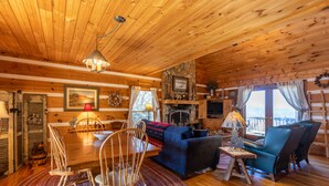 Een knus interieur van een chalet met een houten plafond, een open haard, een eethoek met een tafel en stoelen, en een woonkamer met een bank en fauteuils.