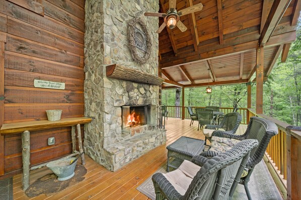 A stone fireplace with a fire, wicker furniture, and a wooden deck with a view of trees.