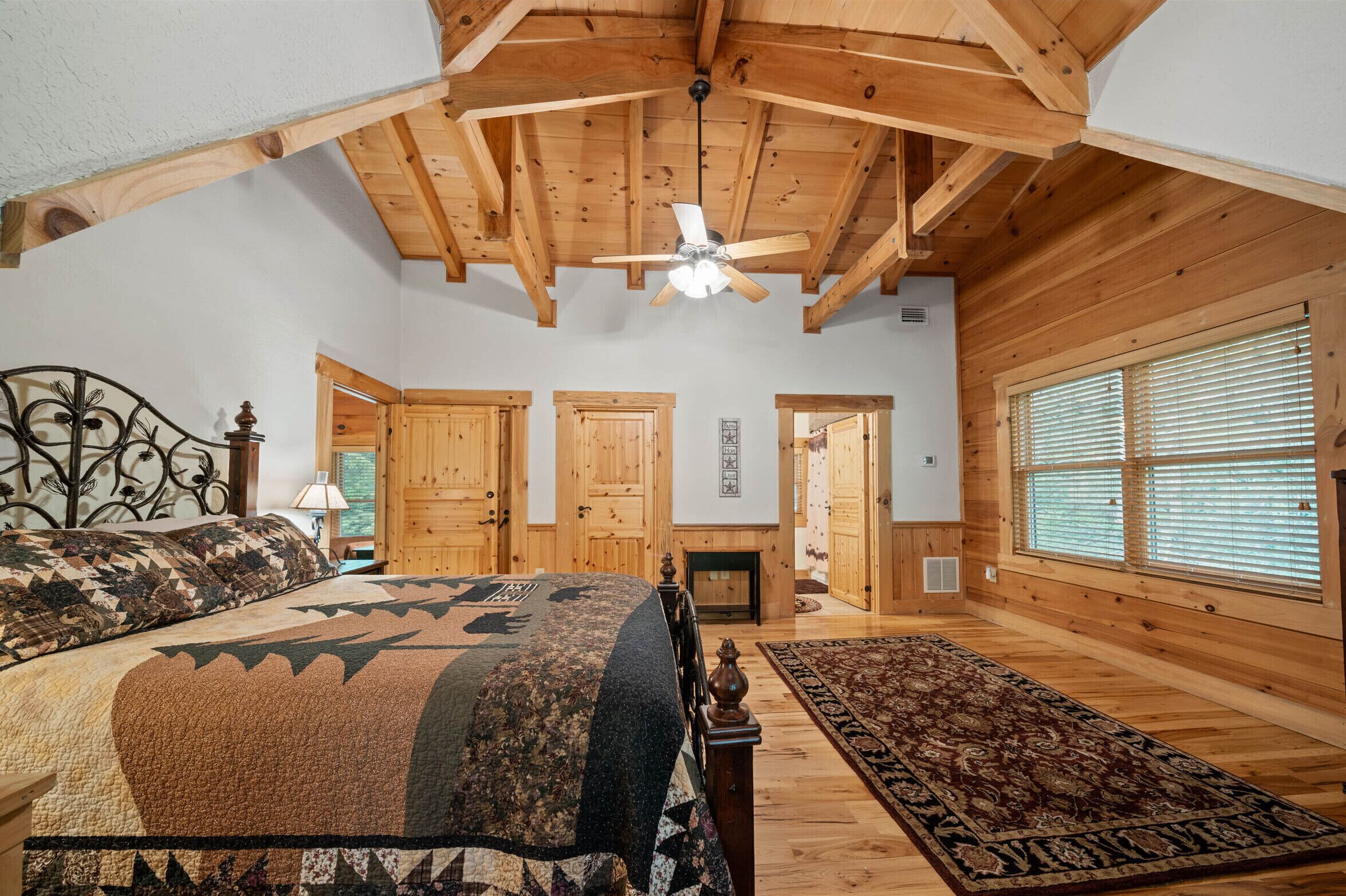 A bedroom with a large bed, wooden ceiling beams, a ceiling fan, and a view of the outside through a window with blinds.