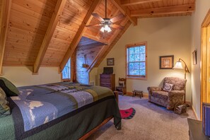 A bedroom with a wooden ceiling, a bed with a quilt, a chair, a desk, and a window with a view.