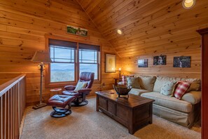 A cozy cabin interior with a wooden ceiling, a leather recliner, a sofa, a coffee table, and a view through the window.
