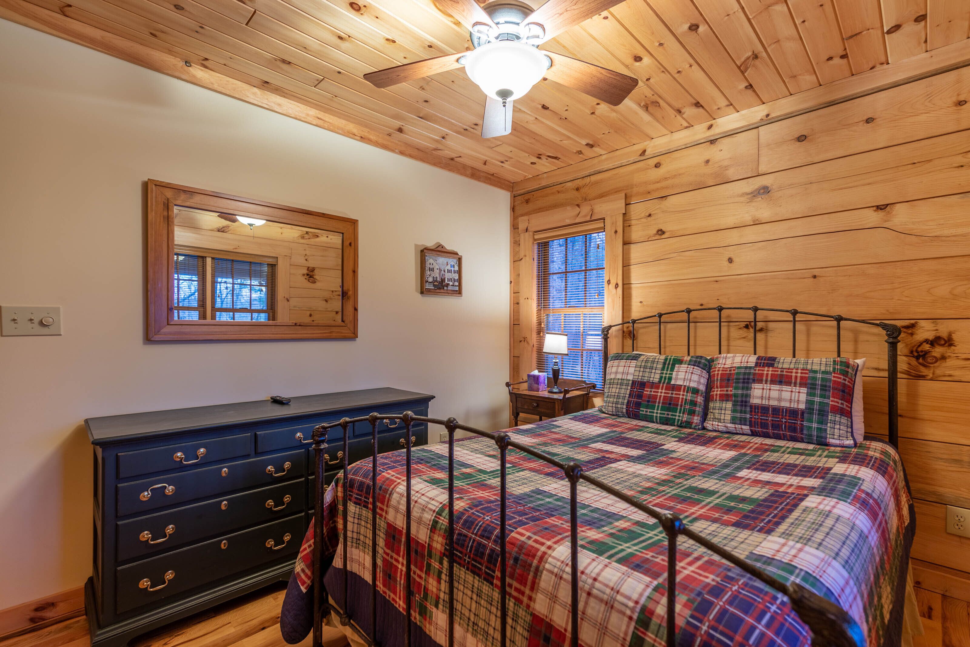 A bedroom with a wooden ceiling, a bed with a plaid bedspread, a dresser with drawers, and a window with blinds.