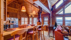 A log cabin interior with a wooden dining area, bar stools, a sofa, and a large window overlooking a mountainous landscape. - Four points, mountain views, king with fireplace on main, hot tub, pet friendly (Vilas)