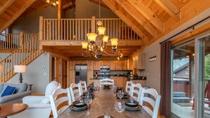 Un comedor amplio con una larga mesa de madera dispuesta para seis personas, rodeada de sillas blancas. Un candelabro cuelga del techo, y la habitación cuenta con vigas de madera y vistas a una terraza exterior a través de puertas de vidrio grandes.
