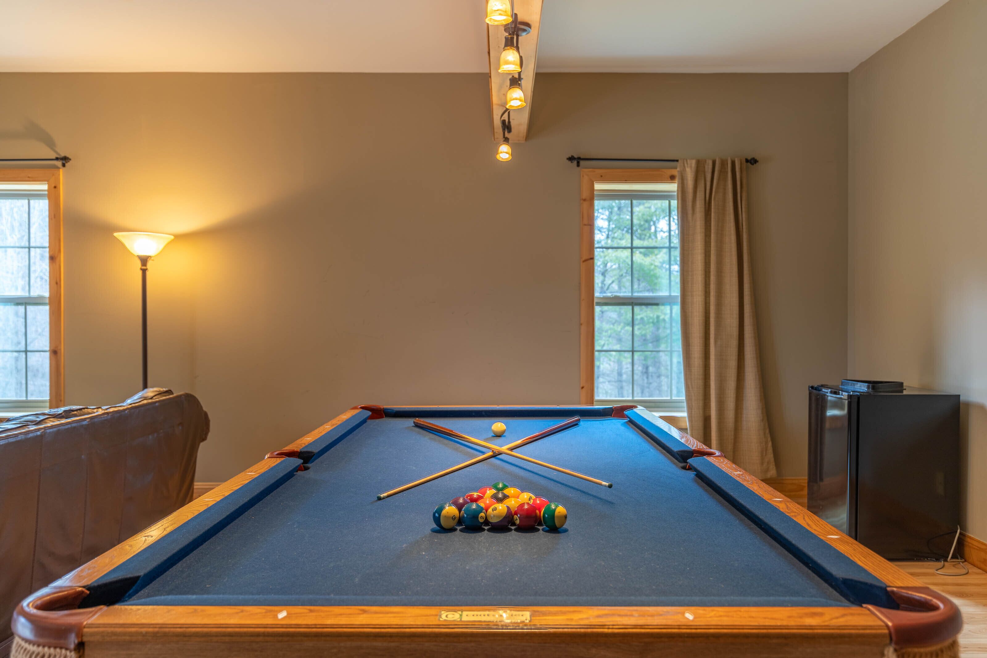 Une table de billard avec des billes et des queues, une lampe et un téléviseur, dans une pièce dotée de fenêtres et de rideaux.