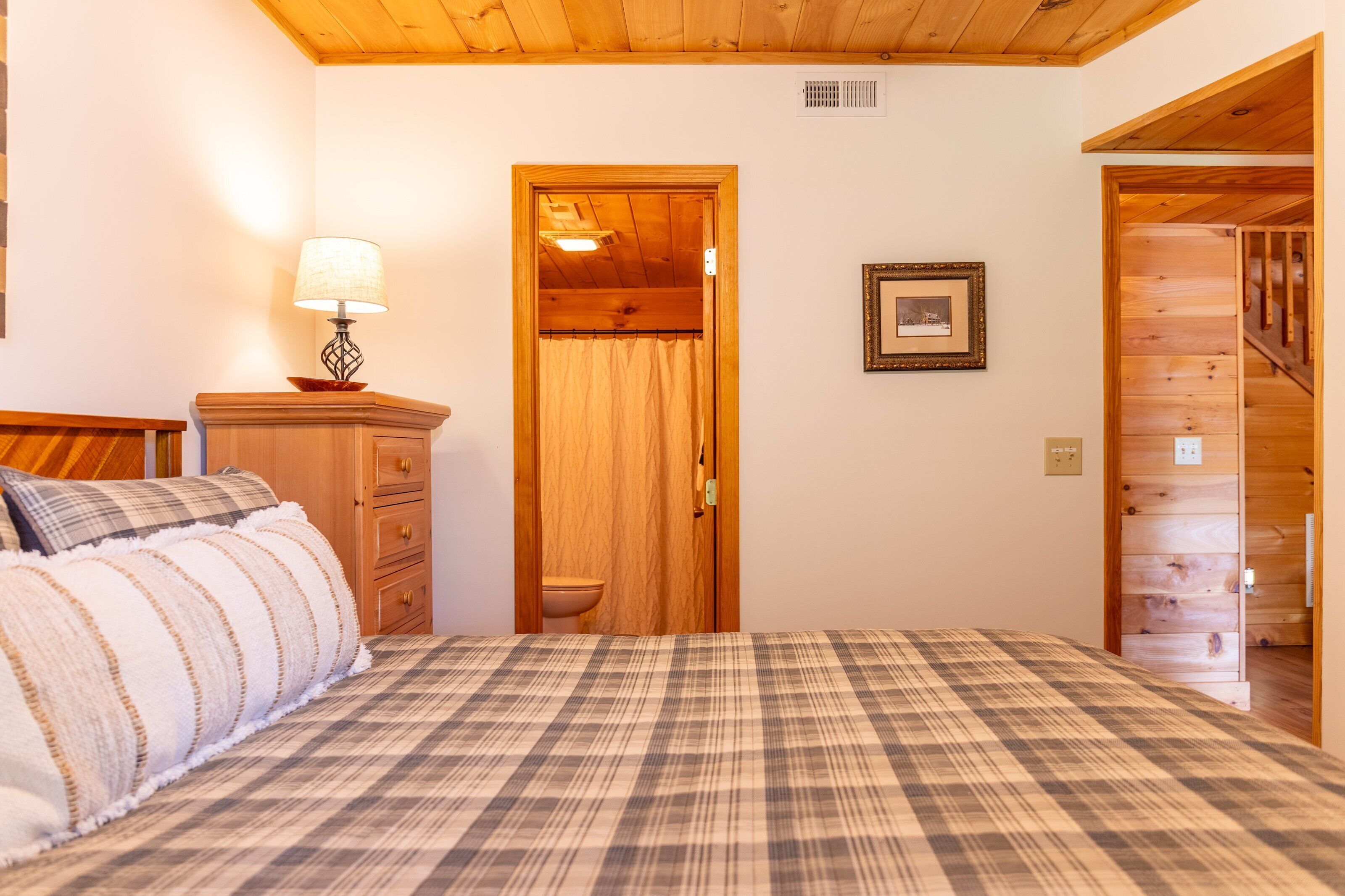 A bedroom with a plaid bedspread, wooden headboard, a nightstand with a lamp, a wooden dresser, a door leading to a bathroom, and a framed picture on the wall.