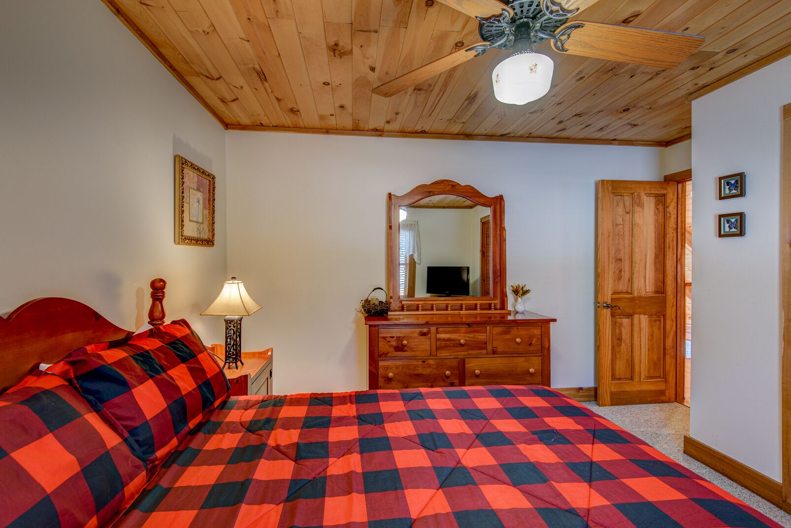 A bedroom with a wooden ceiling, a bed with a red and black checkered blanket, a wooden dresser, a mirror, and a door.
