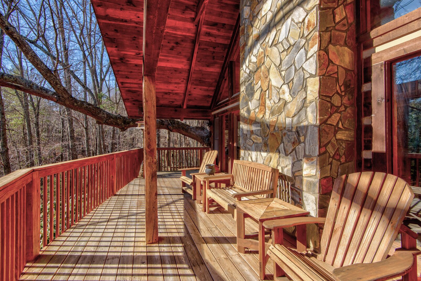A covered wooden deck with stone walls, wooden furniture, and a view of the surrounding forest.