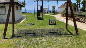 A swing set in a grassy area with a thatched-roof building and a view of the ocean. - Mi Capricho P.5, Apartment with Sea and Pool Views (Mijas)