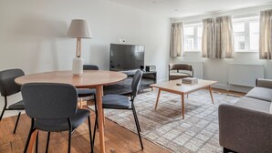 A modern living room with a round wooden table, grey upholstered chairs, a flat-screen TV, a small side table with a lamp, and a sofa. - Deluxe Marylebone 2BR nr Oxford St, by Blueground (London)