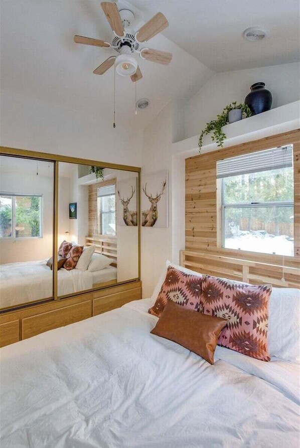 Un dormitorio con cama, un espejo grande, un ventilador de techo, una ventana con persianas y una decoración de cabeza de ciervo.