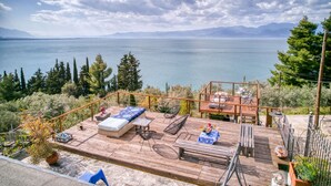 Una terraza de madera con mobiliario exterior, una mesa con mantel azul y vistas a un lago y montañas.