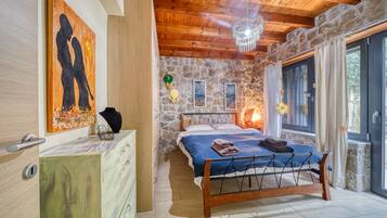 Een slaapkamer met een stenen muur, houten plafond, een bed met metalen frame, een kroonluchter en kunstwerken aan de muur.