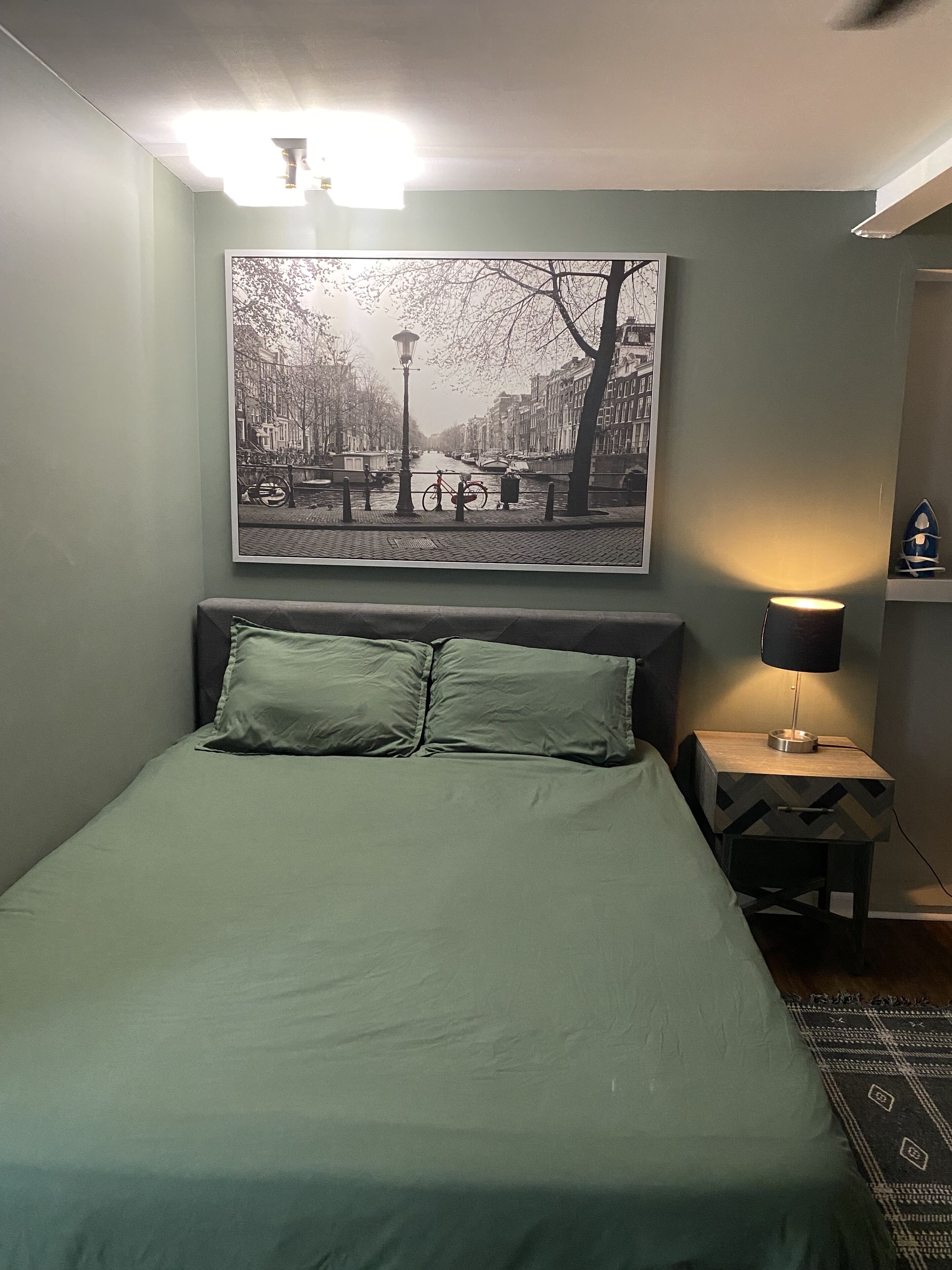 Ein Schlafzimmer mit einem Bett, einem Nachttisch, einer Lampe und einem an der Wand hängenden Stadtbild.