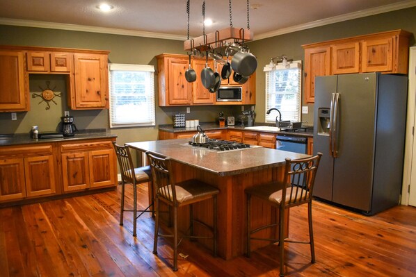 A kitchen with wooden cabinets, a stainless steel refrigerator, and a central island with bar stools. - Youngsville Retreat- pool, private pond, outdoor fireplace (Broussard)