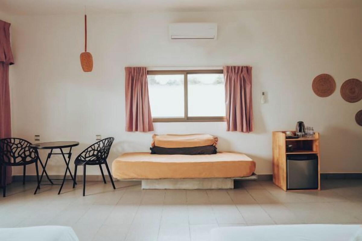 A small, well-lit room with a bed, a table with chairs, a small cabinet, and a window with curtains.