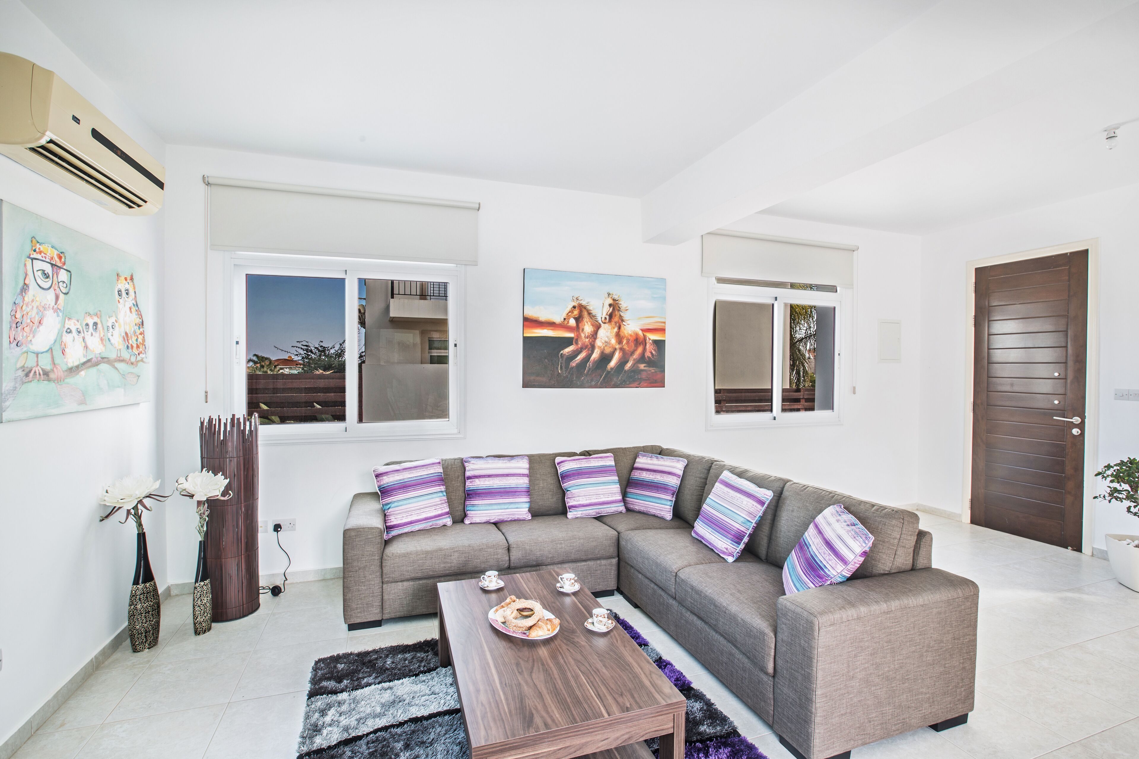 A modern living room with a sectional sofa, a coffee table, and decorative pillows. There are two windows with views of the outside, and a door to the right.