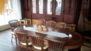 Une table à manger en bois, avec six chaises, un grand saladier rempli de pain au centre, et une fenêtre avec des stores.