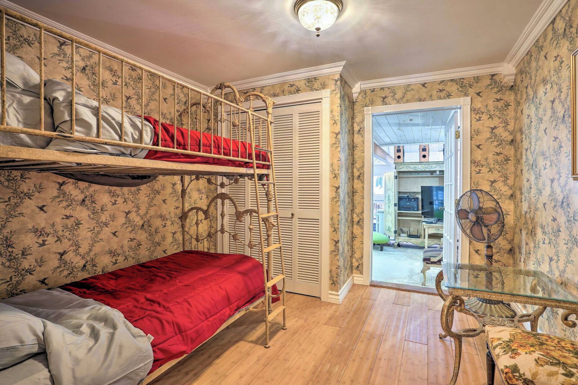 Quarto com beliches, uma mesinha, um ventilador e uma porta que dá para outro espaço.