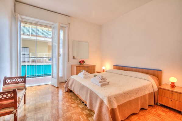 A bedroom with a bed, bedside table, nightstand, and a view of a balcony.