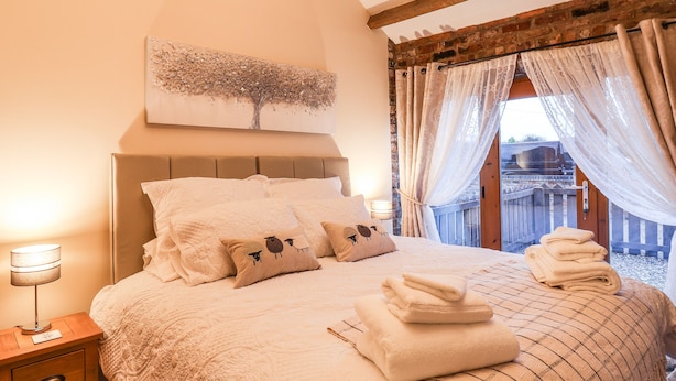 A neatly made bed with a quilt, pillows, and folded towels. There is a bedside table with a lamp and a small drawer. A wall-mounted artwork of a tree is above the bed.