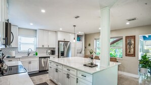 A modern kitchen with white cabinetry, a large island, stainless steel appliances, and a view of greenery outside.