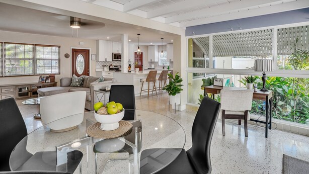 A modern kitchen with white cabinets, a dining area with a glass table, and a view of greenery outside.