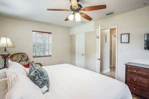 Un dormitorio con cama, ventilador de techo, ventana con persianas, cómoda y puerta que da a otra habitación.
